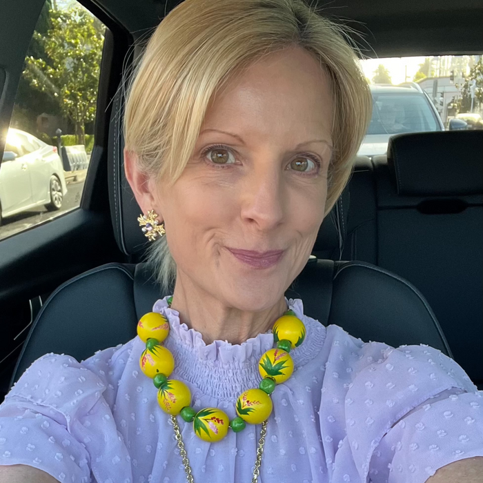 Woman wearing a purple polka dot blouse and yellow beaded necklace sitting in a car.