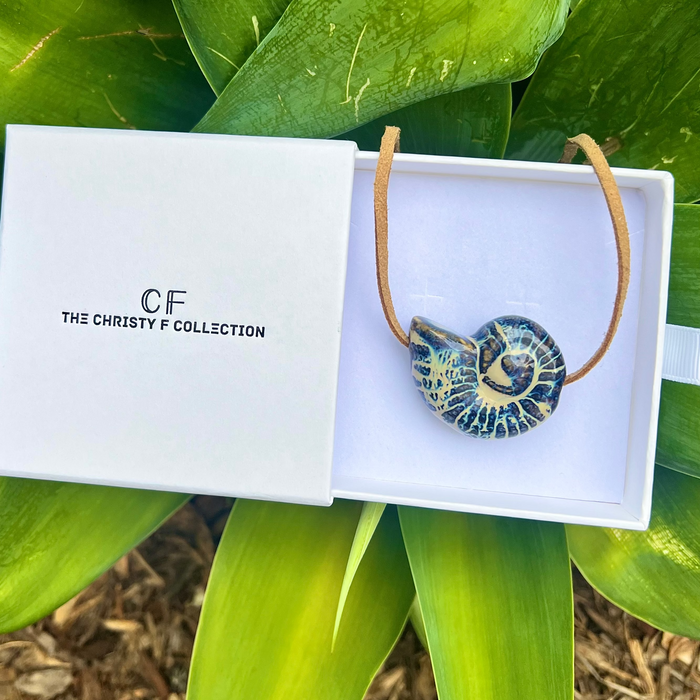 Necklace with a decorative pendant in a 'The Christy F Collection' box, surrounded by green leaves.