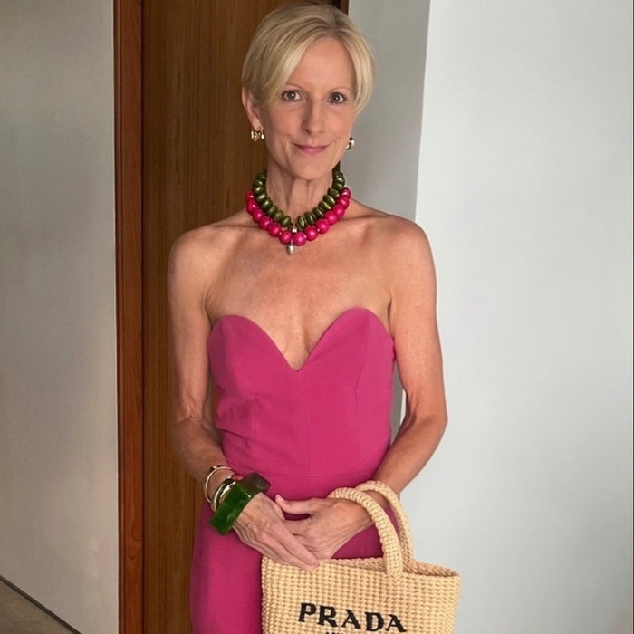 A woman wearing a bright pink strapless dress and a chunky green and pink beaded necklace, holding a beige woven Prada Milano tote bag. She is standing indoors against a white wall and a wooden door frame.