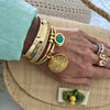 A close-up of a hand wearing multiple gold bracelets and rings, resting on a woven straw hat. The bracelets include a thick textured gold cuff, a bangle with a green stone, and a round sunburst charm.