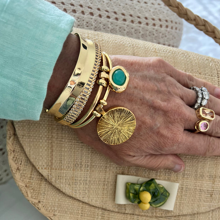 A close-up of a hand wearing multiple gold bracelets and rings, resting on a woven straw hat. The bracelets include a thick textured gold cuff, a bangle with a green stone, and a round sunburst charm.