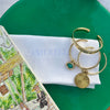 A close-up styled flat-lay showing a white Camerlie napkin with a gold-tone cuff bracelet and a small green gemstone charm, placed on a green plate next to a printed magazine.