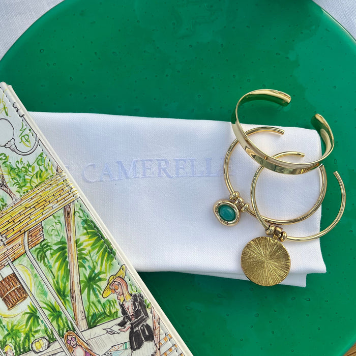 A close-up styled flat-lay showing a white Camerlie napkin with a gold-tone cuff bracelet and a small green gemstone charm, placed on a green plate next to a printed magazine.