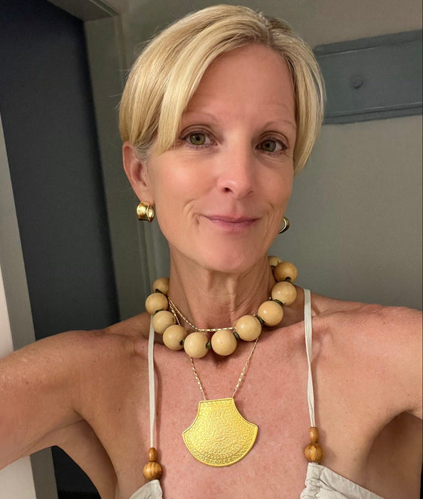 A woman with short blonde hair wearing a gold pendant necklace with a scalloped edge, a beaded wooden necklace, and matching gold earrings, posing in a light-colored halter top.