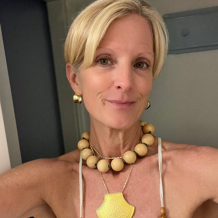 A woman with short blonde hair wearing a gold pendant necklace, a beaded wooden necklace, and matching gold earrings, paired with a light-colored spaghetti strap top.
