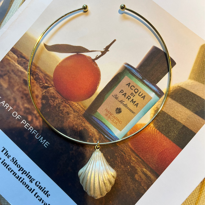 A gold-tone circular hoop earring with a dangling textured shell charm, displayed over a perfume magazine page featuring an orange and an Acqua Parma bottle.