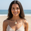 A woman with long dark brown hair smiling on a sandy beach, wearing a delicate gold chain necklace with a white seashell pendant and a beige spaghetti-strap top.