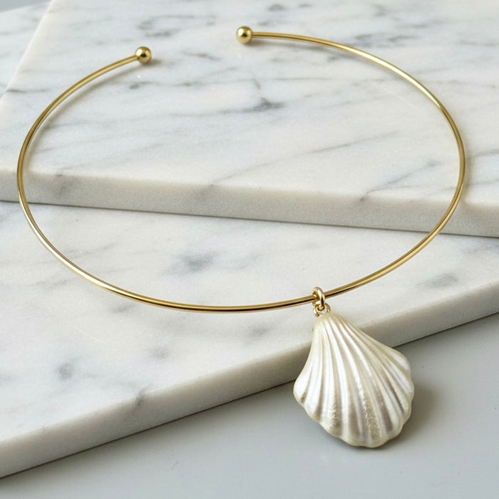 A delicate gold wire bangle necklace with a textured white shell pendant, displayed on a white marble background.