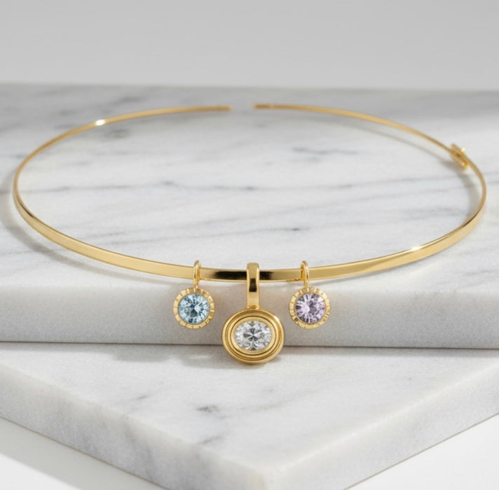 A gold necklace with three hanging charms, including a central round bezel-set clear stone and two smaller round stones, displayed on a white marble block.