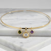 A gold necklace with three hanging charms, including a central round bezel-set clear stone and two smaller round green and purple stones, displayed on a white marble block.