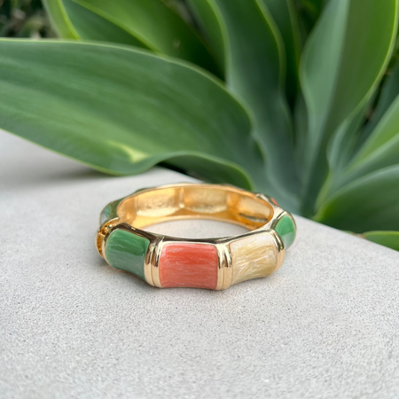 Gold bangle with green and pink inlay on a white surface with green leaves in the background