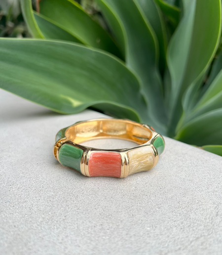 Gold bangle with green and pink inlay on a white surface with green leaves in the background