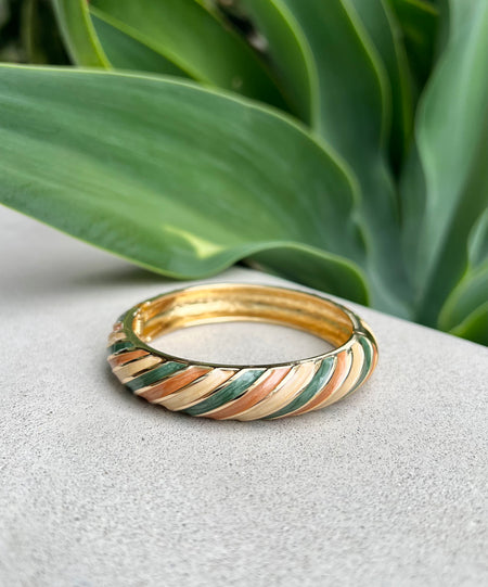 Gold and silver bangle with a striped pattern on a white surface with green leaves in the background