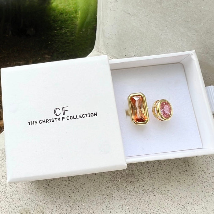 A pair of gold-toned statement ring displayed in an open white jewelry box. One stone has a rectangular faceted amber-orange stone, and the other has a round pink stone, both set in gold.