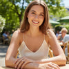 A smiling woman with brown hair sits at an outdoor cafe table wearing a cream-colored spaghetti-strap camisole top and a ring with a pink gemstone.