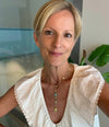A woman with short blonde hair wearing a white textured blouse and a gold pendant necklace with blue and green stones, photographed indoors with a glass wall and plant in the background.