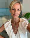 A woman with short blonde hair wearing a white textured blouse and a gold pendant necklace with blue and green stones, photographed indoors with a glass wall and plant in the background.