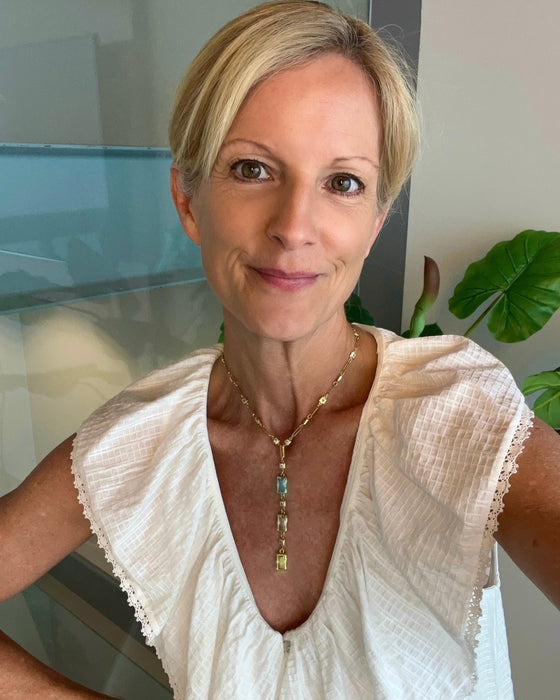 A woman with short blonde hair wearing a white textured blouse and a gold pendant necklace with blue and green stones, photographed indoors with a glass wall and plant in the background.