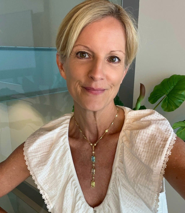 A woman with short blonde hair wearing a white textured blouse and a gold pendant necklace with blue and green stones, photographed indoors with a glass wall and plant in the background.