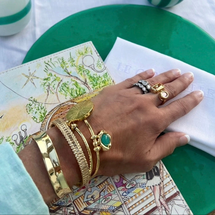 A close-up of a hand wearing multiple gold bracelets and rings, resting on a green plate over a patterned book and white napkin.