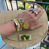 A close-up lifestyle shot of a person's hand resting on a woven straw clutch bag. The hand is adorned with multiple gold bracelets, rings, and a turquoise stone earring.