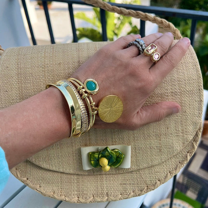 A close-up lifestyle shot of a person's hand resting on a woven straw clutch bag. The hand is adorned with multiple gold bracelets, rings, and a turquoise stone earring.