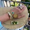 A close-up lifestyle shot of a person's hand resting on a woven straw clutch bag. The hand is adorned with multiple gold bracelets, rings, and a turquoise stone earring.