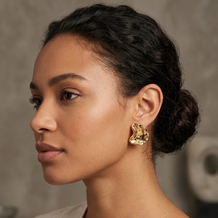 A close-up side profile of a woman with dark hair in a low bun, wearing a textured, organic-shaped gold earring against a soft gray background.