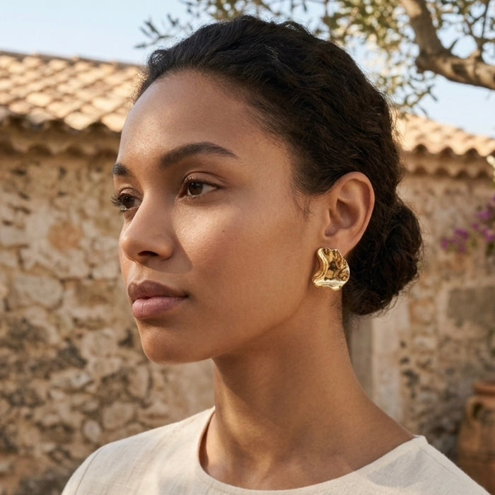 A woman in profile wearing a chunky, organic-shaped gold earring, photographed outdoors against a rustic stone wall and tiled roof.