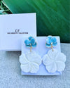 Pair of earrings with blue floral tops and white leaf-like bottoms on a branded display card.
