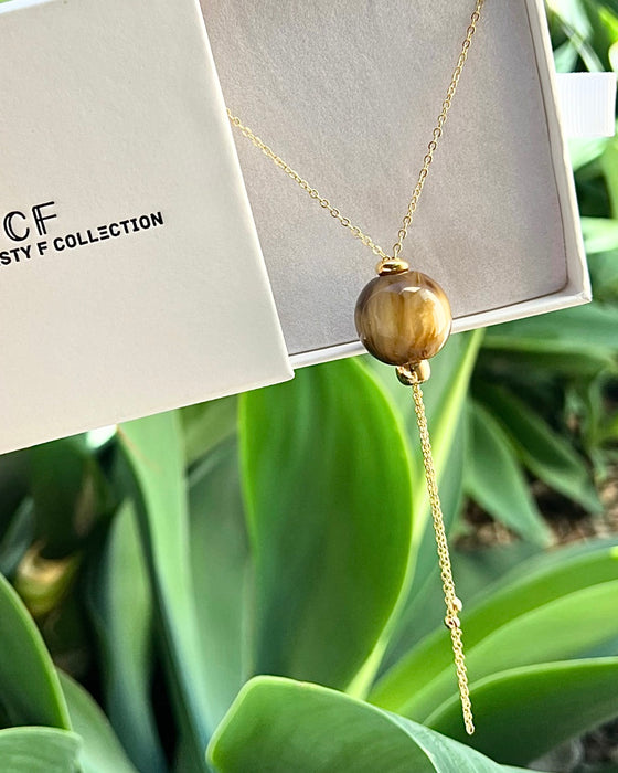 Gold necklace with a large brown bead on a white card with 'CF' branding, set against green leaves.