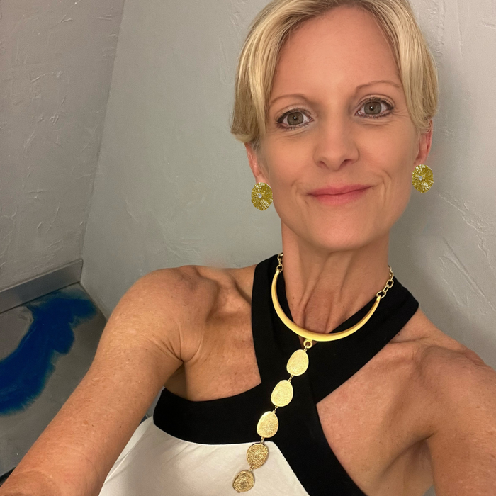 A woman wearing a gold disc pendant necklace and matching gold earrings, styled with a black and white halter top, taking a mirror selfie.