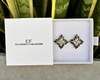 Pair of earrings in a white box with 'The Christy F Collection' branding, against a plant background.