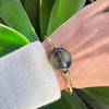 Hand wearing a gold bracelet with a large blue stone, against a green leafy background