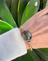 Hand wearing a gold bracelet with a large blue stone, against a green leafy background