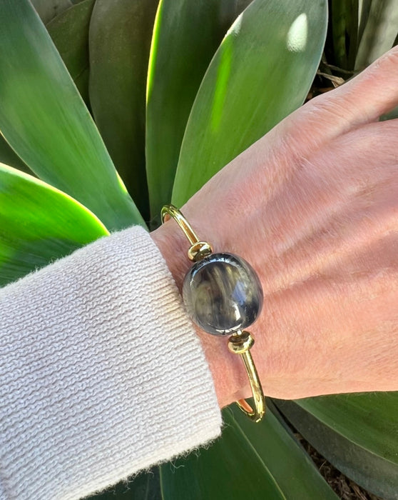 Hand wearing a gold bracelet with a large blue stone, against a green leafy background