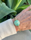 Hand wearing a gold bracelet with a large turquoise bead against a green leafy background