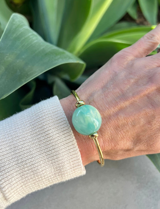 Hand wearing a gold bracelet with a large turquoise bead against a green leafy background
