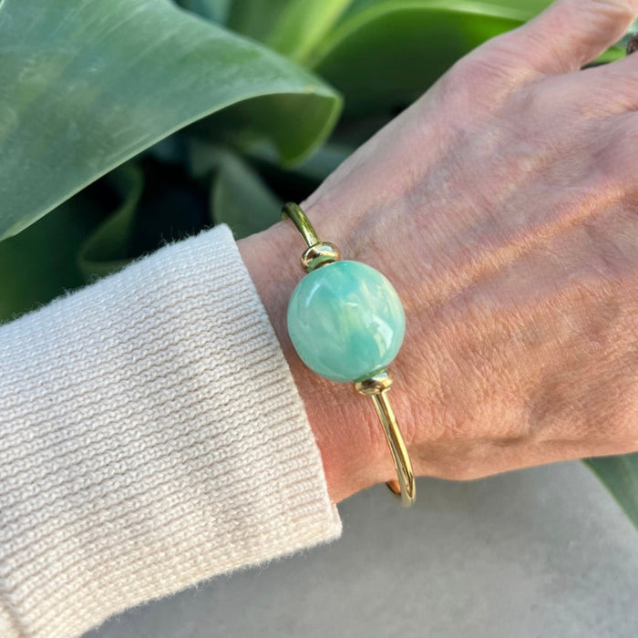 Hand wearing a gold bracelet with a large turquoise bead against a green leafy background