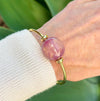 Gold bracelet with a pink bead on a wrist against a green leafy background