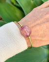 Gold bracelet with a pink bead on a wrist against a green leafy background