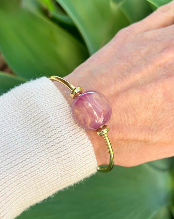 Gold bracelet with a pink bead on a wrist against a green leafy background