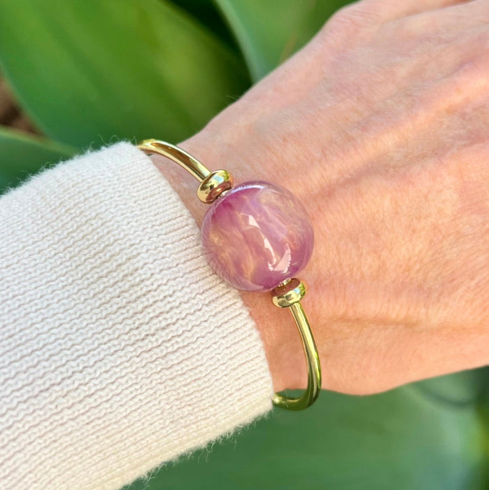 Gold bracelet with a pink bead on a wrist against a green leafy background