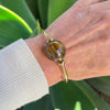 Gold bracelet with a large round gemstone on a wrist against a green leafy background