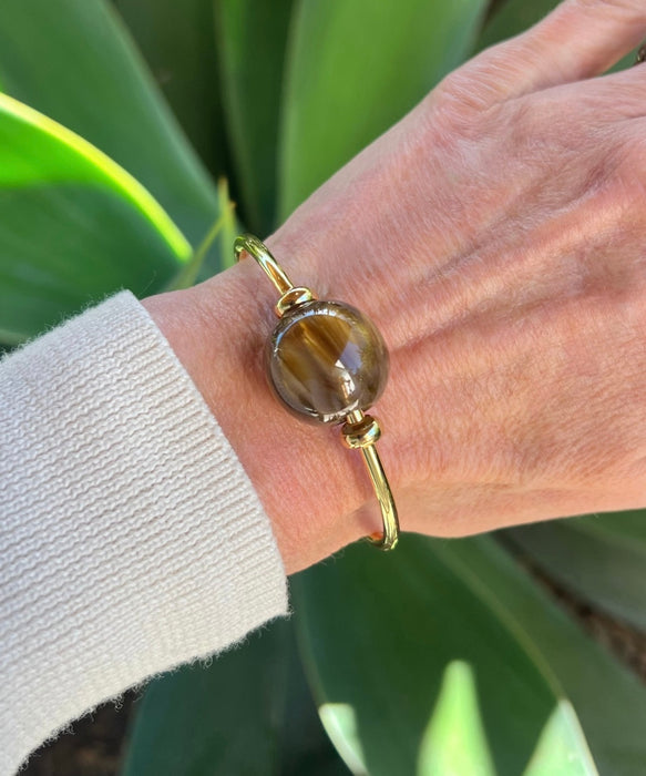 Gold bracelet with a large round gemstone on a wrist against a green leafy background