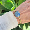 Hand wearing a gold bracelet with a blue bead against a green leafy background