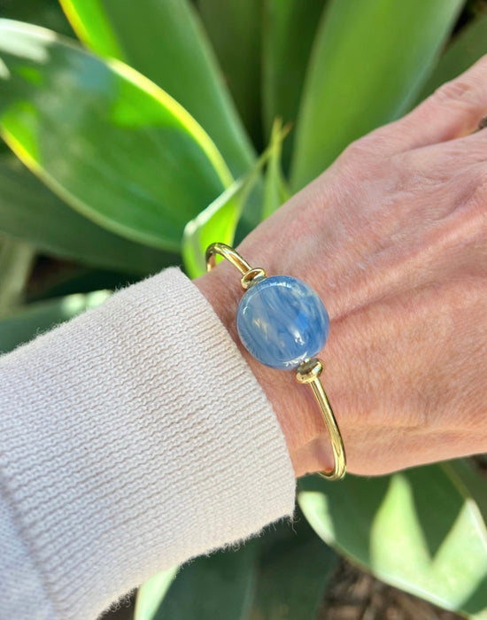 Hand wearing a gold bracelet with a blue bead against a green leafy background