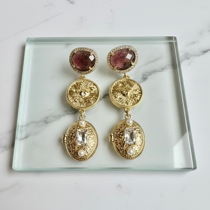 Gold earrings with gemstones on a clear glass tray against a marble background