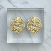 Gold earrings with diamond centers on a clear stand against a marble background