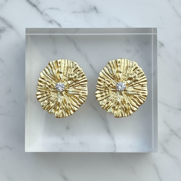 Gold earrings with diamond centers on a clear stand against a marble background
